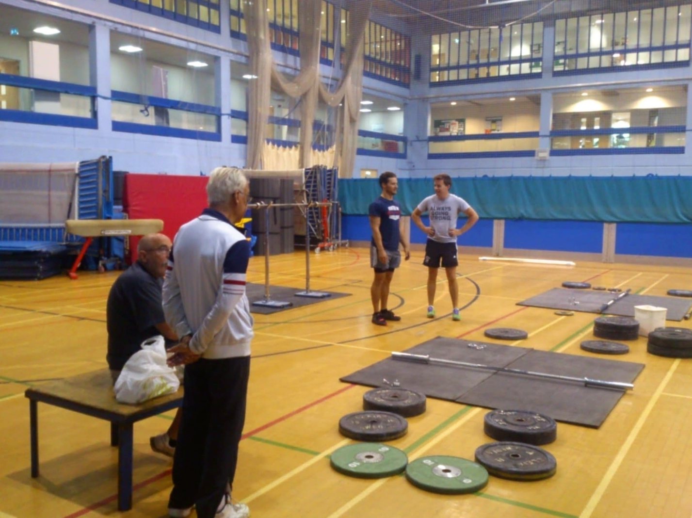 Brian in a tracksuit watching two lifters by the platforms in a sports hall