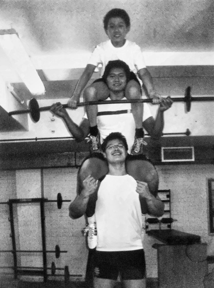 Three Chea brothers stacked on each other's shoulders in the club gym, the youngest holding a barbell